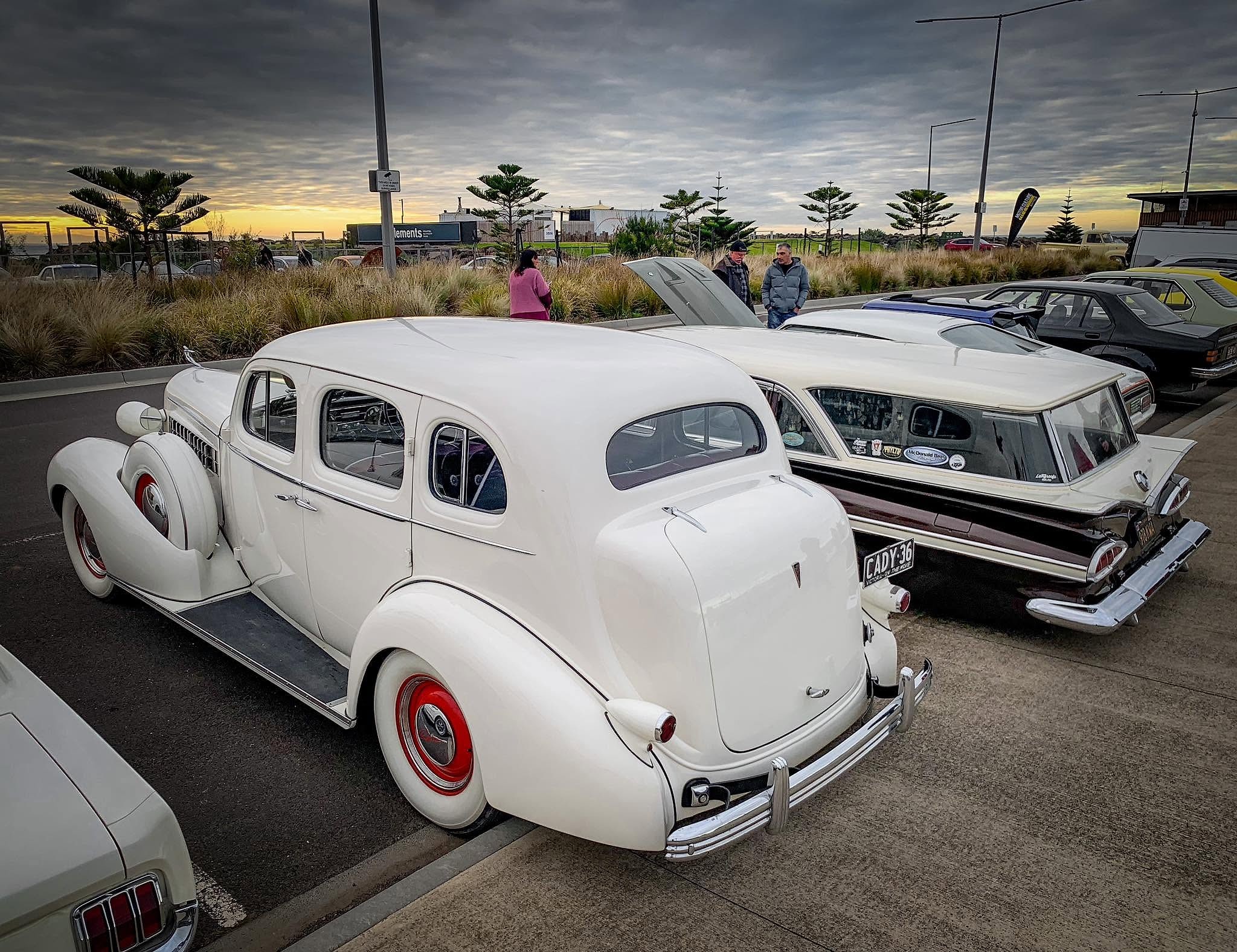 A white 1936 Cadillac with red steel wheels and whitewall tyres parked at a car show at sunset, beside a two tone American wagon