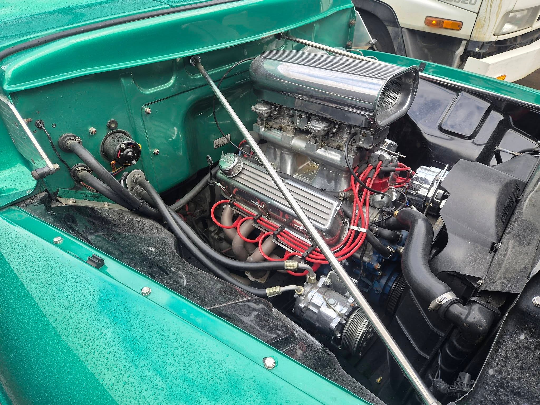 A blown V8 sitting in the teal engine bay of a Ford F100, with braided lines and a polished blower drive