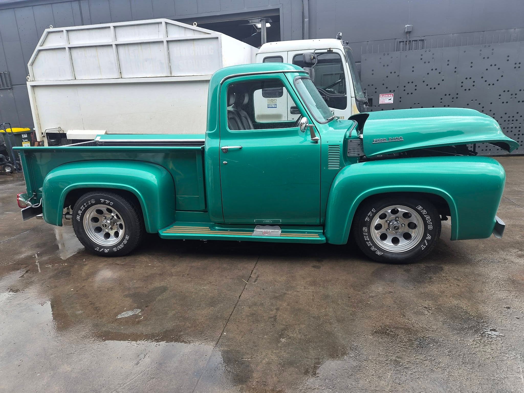 A teal Ford F100 pickup with a white canopy over the tray, parked on wet concrete outside the workshop