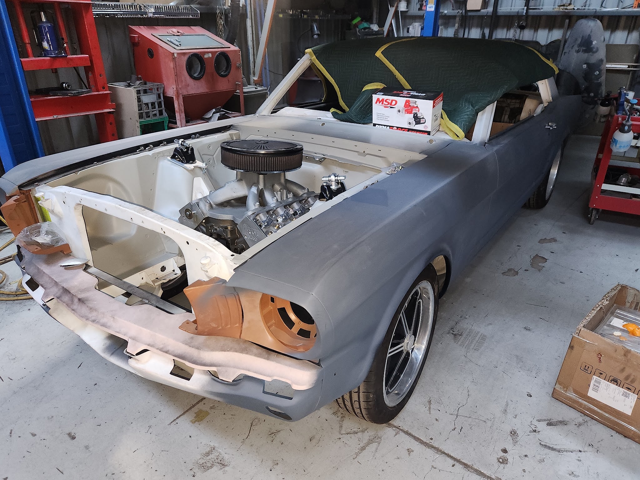 A first generation Mustang convertible part way through a restoration, one guard in grey primer, the boot lid off and a fresh V8 fitted