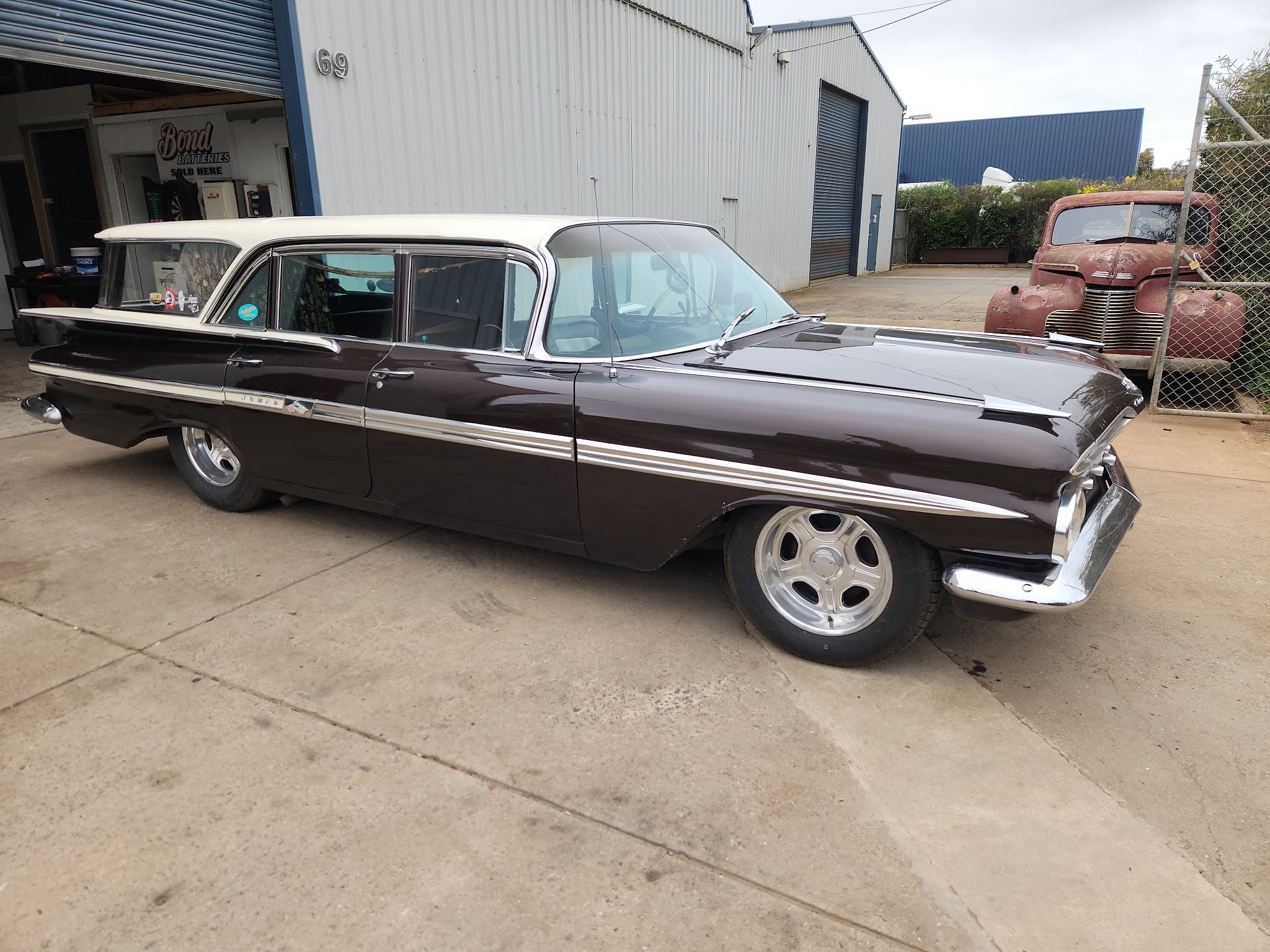 A long American wagon in maroon over cream outside the workshop with a rusted 1940s coupe behind the fence
